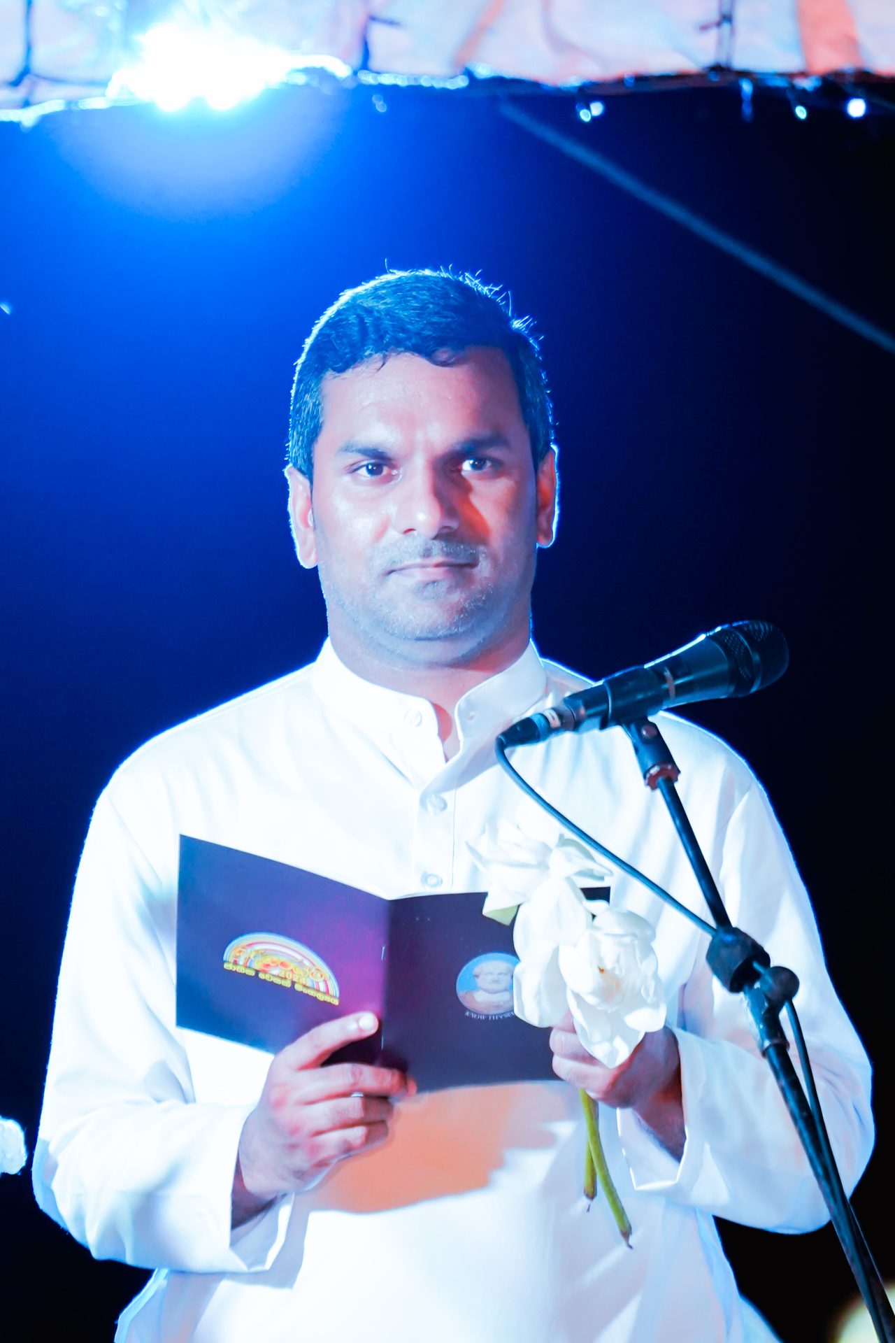 Man delivering speech holding flowers and booklet under blue light.