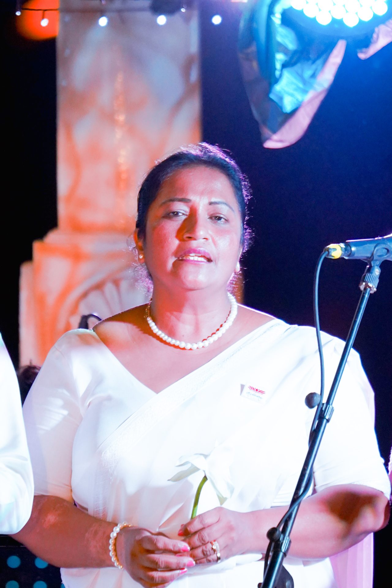 Woman in white saree speaking at formal event.