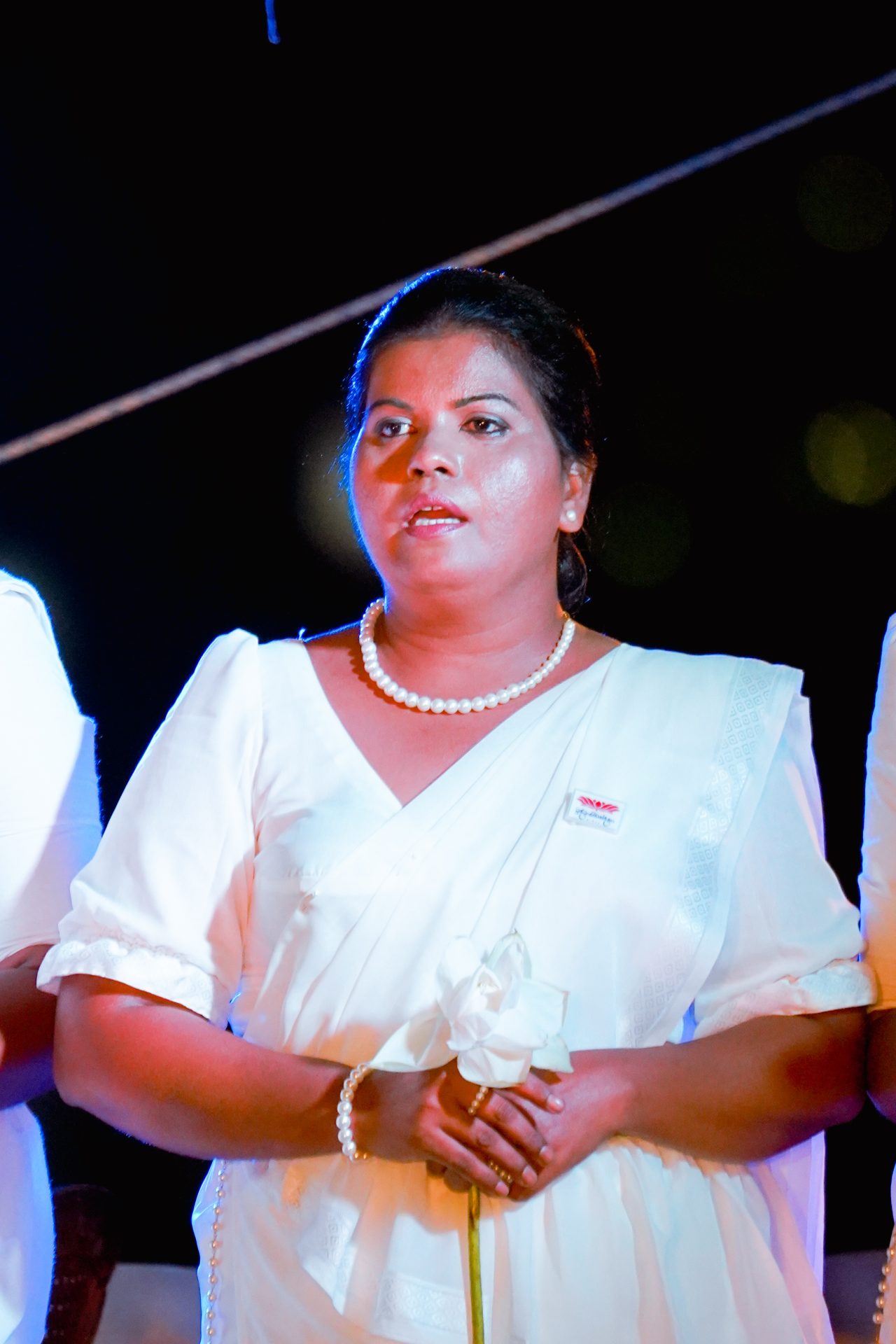 Woman in white saree holding a lotus flower at a ceremonial event.