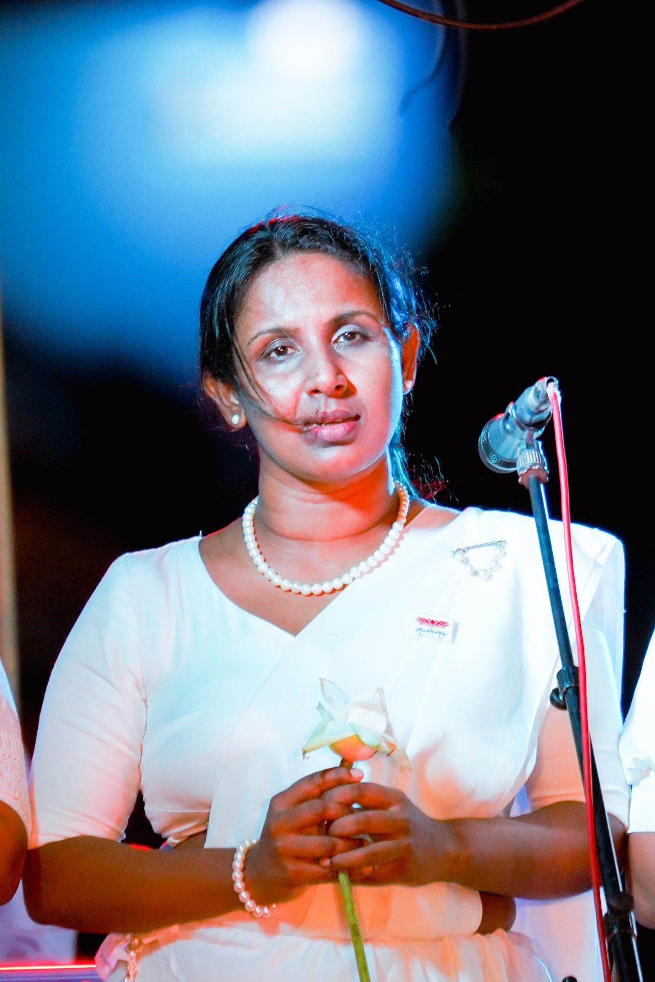 Woman in white sari holding lotus speaks into microphone.