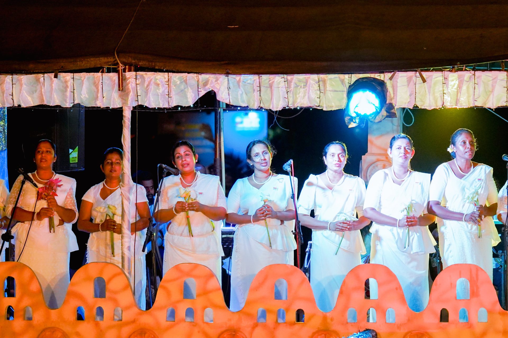 Women in white dresses holding flowers on stage during a ceremonial event.