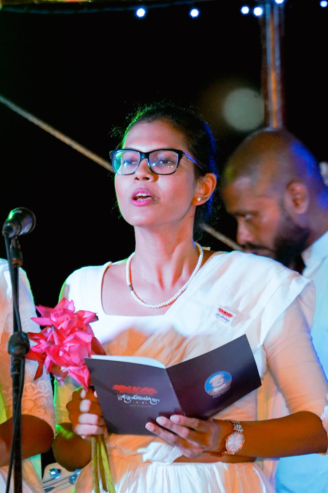 Woman in saree speaking at a formal event, holding flowers and a booklet.