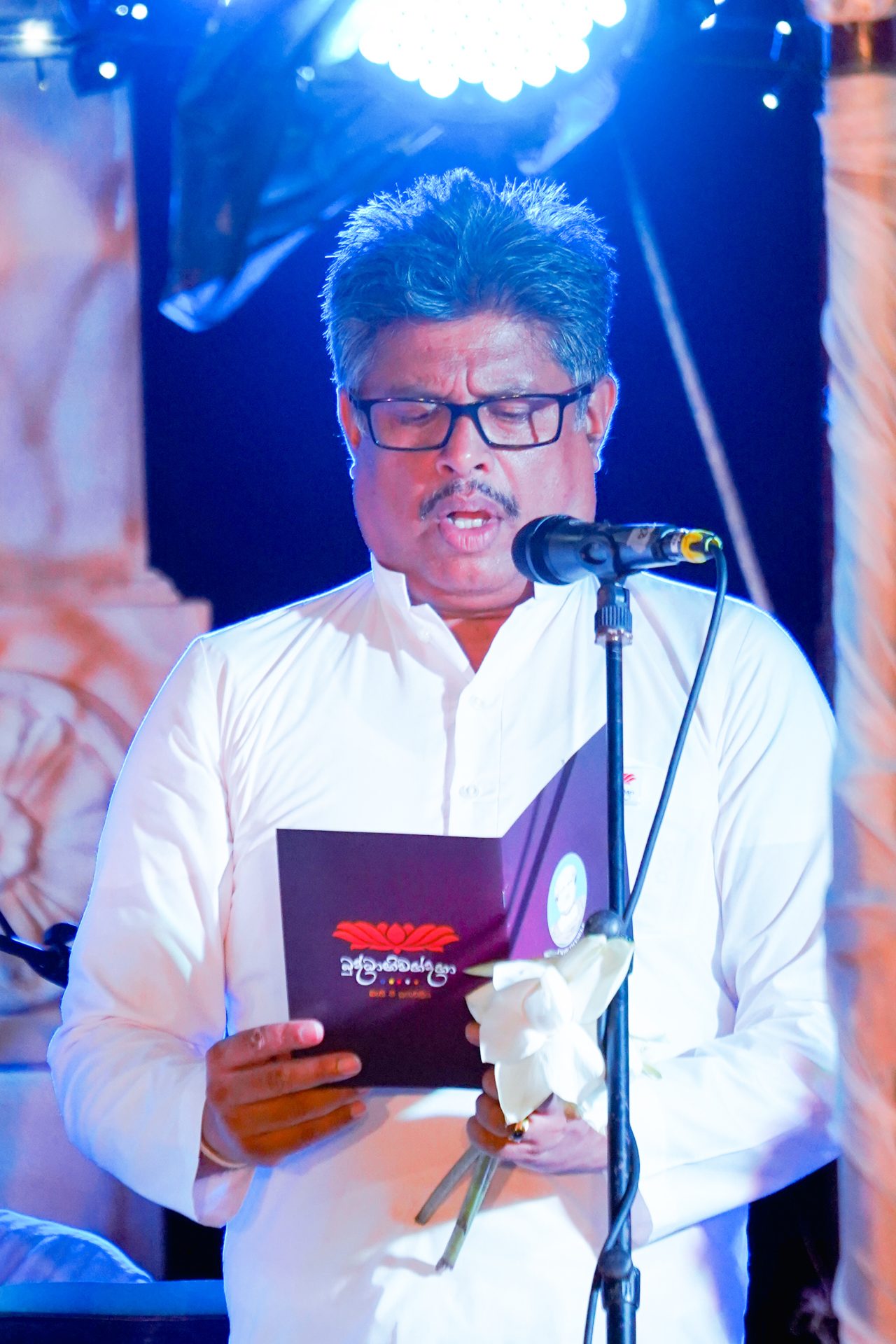 Middle-aged man speaking at formal event, holding flower and booklet under dramatic lighting.