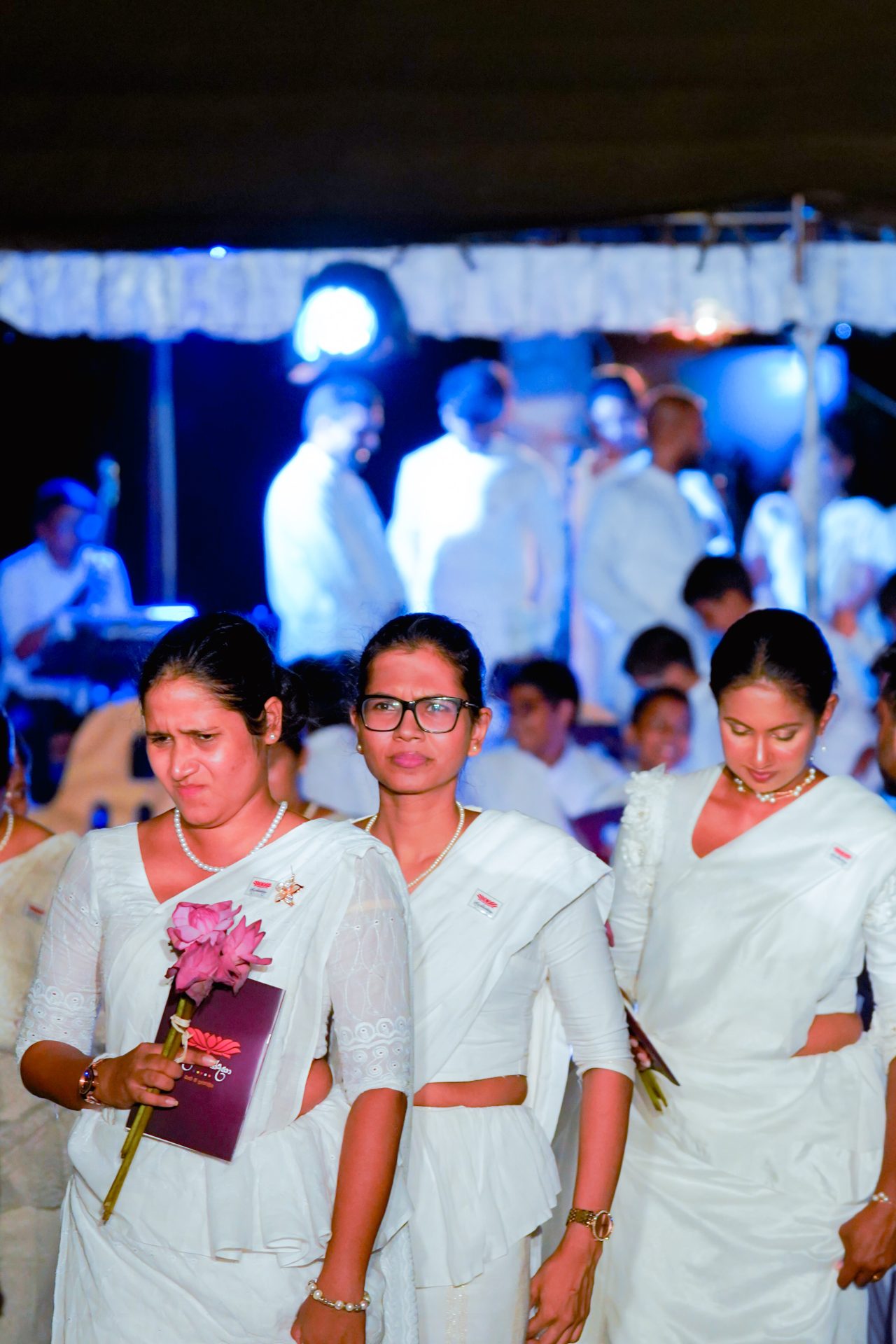 Women in white sarees at a solemn event holding a lotus flower.