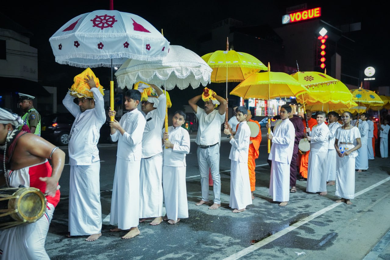 Nighttime religious procession with traditional attire, ornate umbrellas, and rhythmic drumming on urban street.