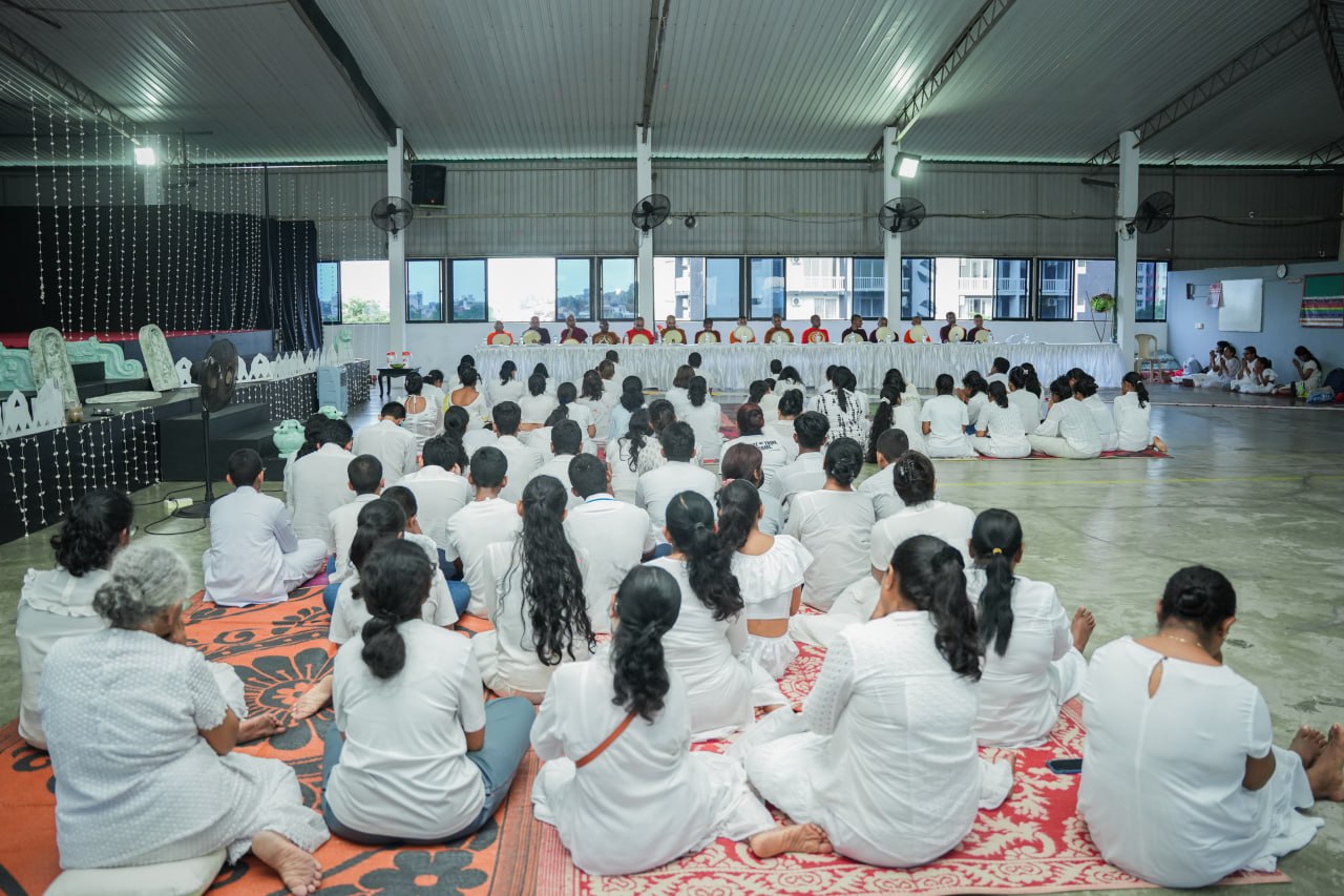 Large group meditation in well-lit hall with white-dressed attendees.
