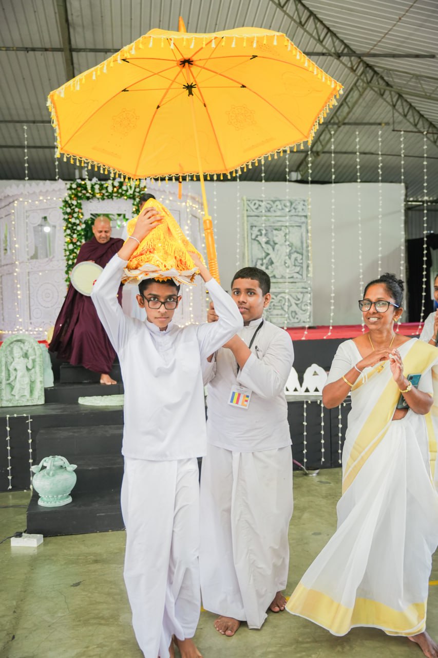 Ceremonial event with boys in white, traditional attire, under parasol, woman clapping, monk playing drum.