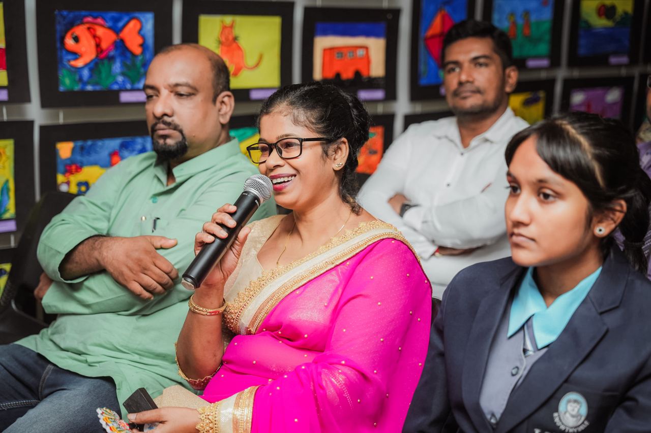 Community Art Event: Speaker Engages with Audience Woman in pink saree speaks at art exhibition, audience engaged.
