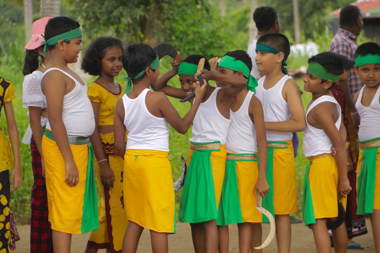 Children in yellow costumes joyfully perform a cultural dance outdoors amidst lush greenery.