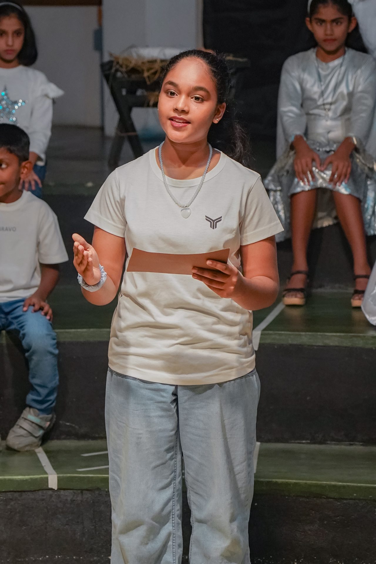 Girl leads school play performance, confidently delivering speech with peers in themed setting.