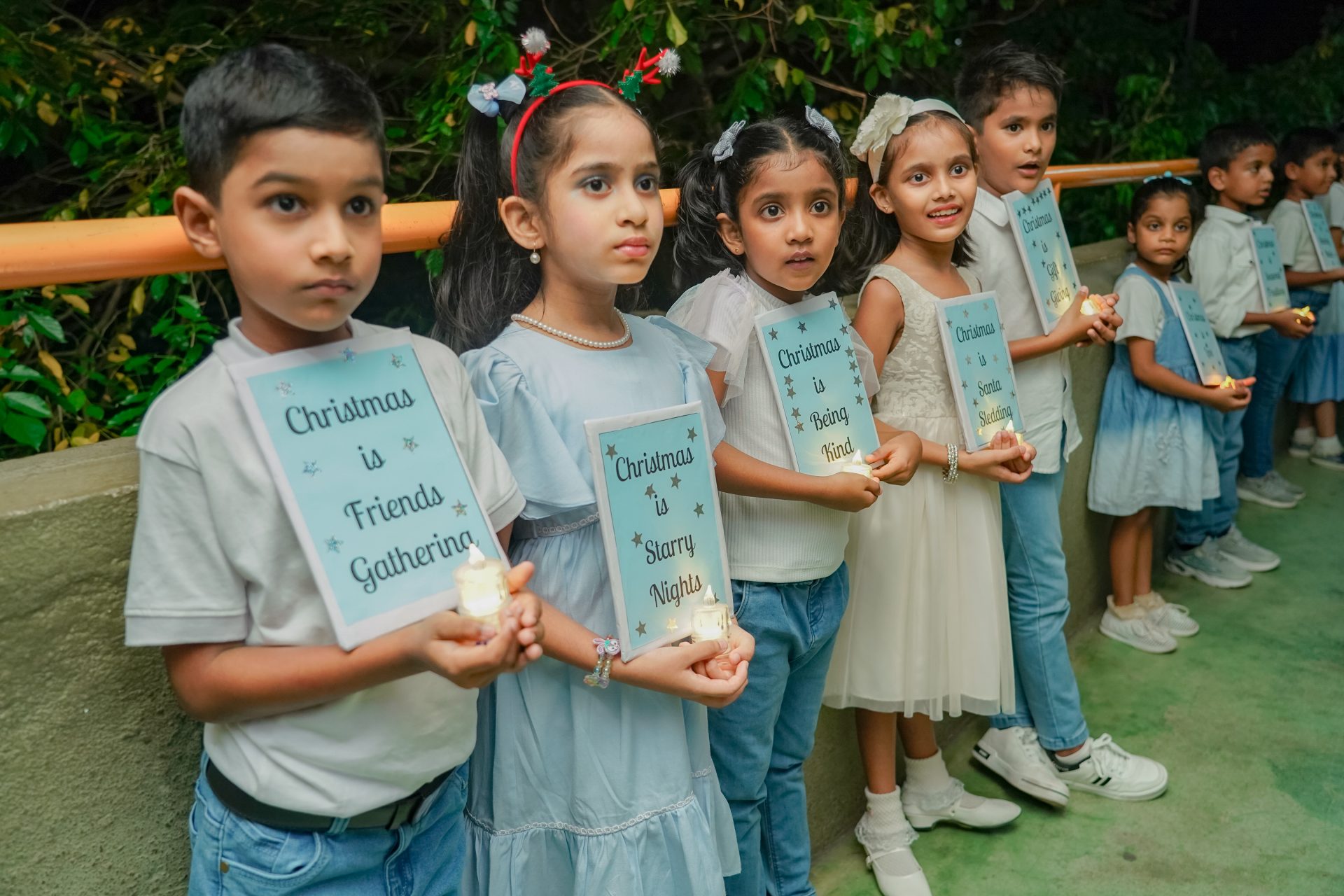 Children in light blue uniforms hold candles during a solemn ceremony in the park.