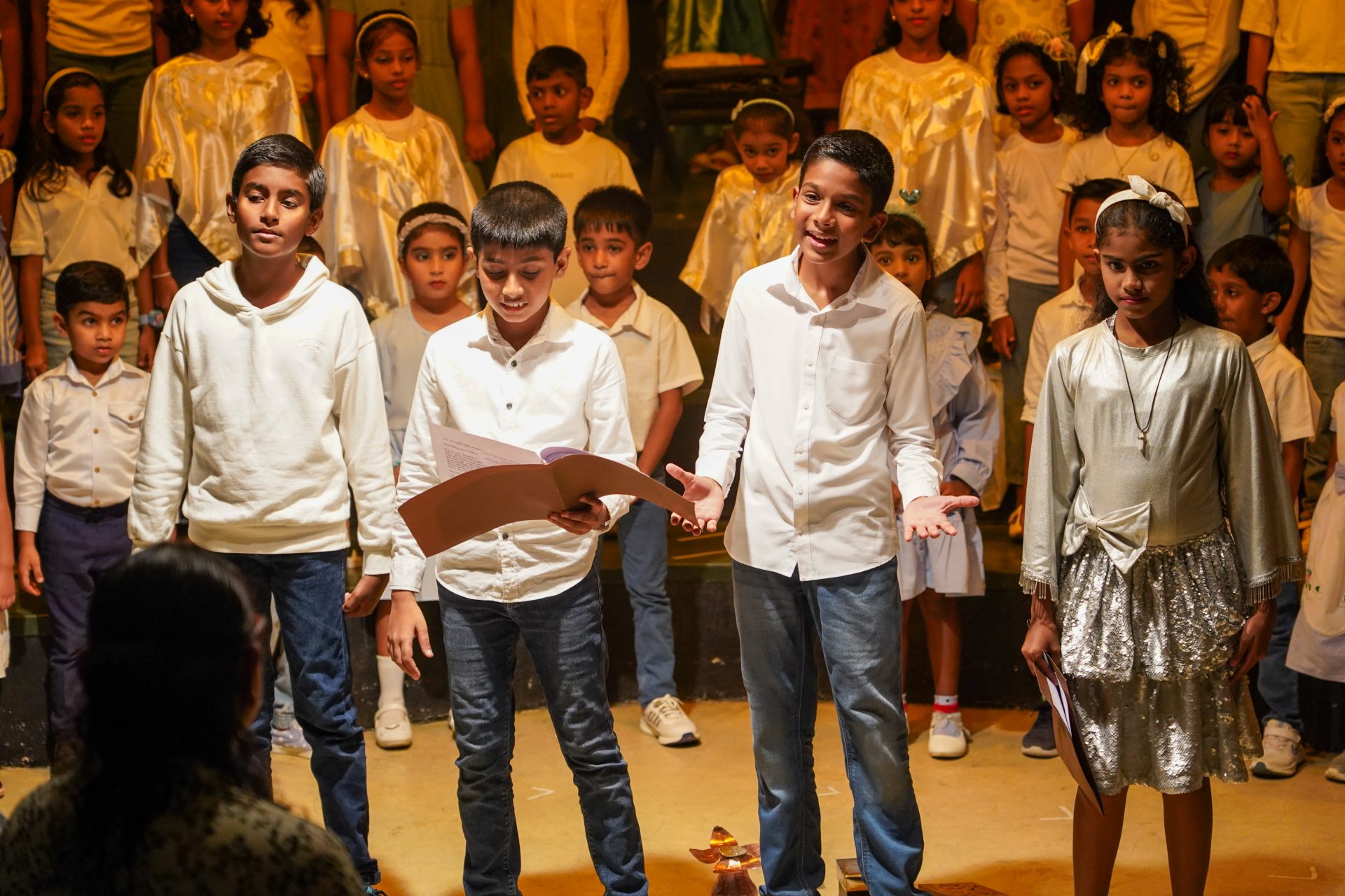 Children perform in school play; boys in white, girl in silver, choir in background.