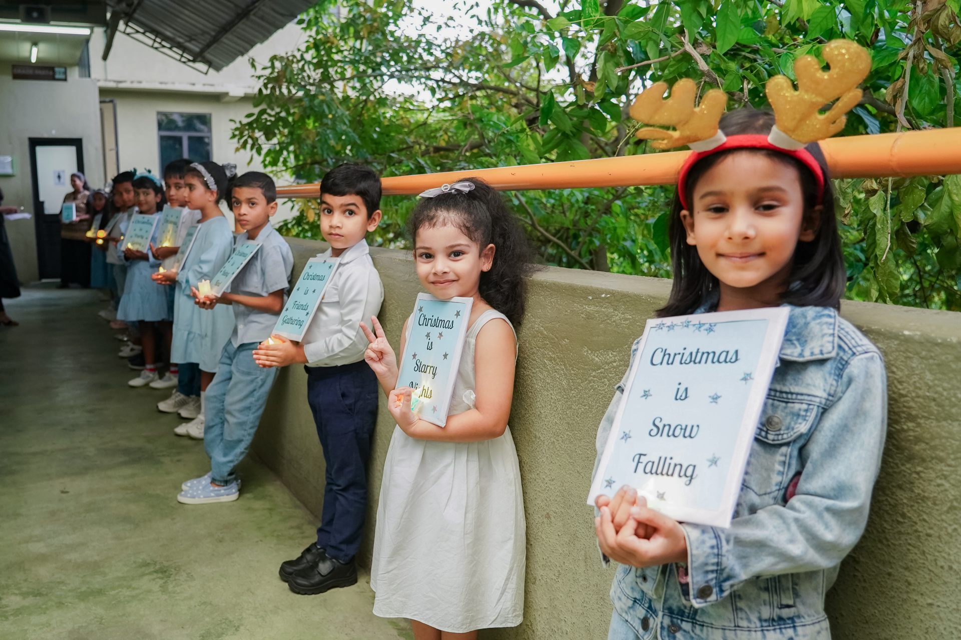 Children in festive attire hold holiday-themed papers in a sunlit school corridor.