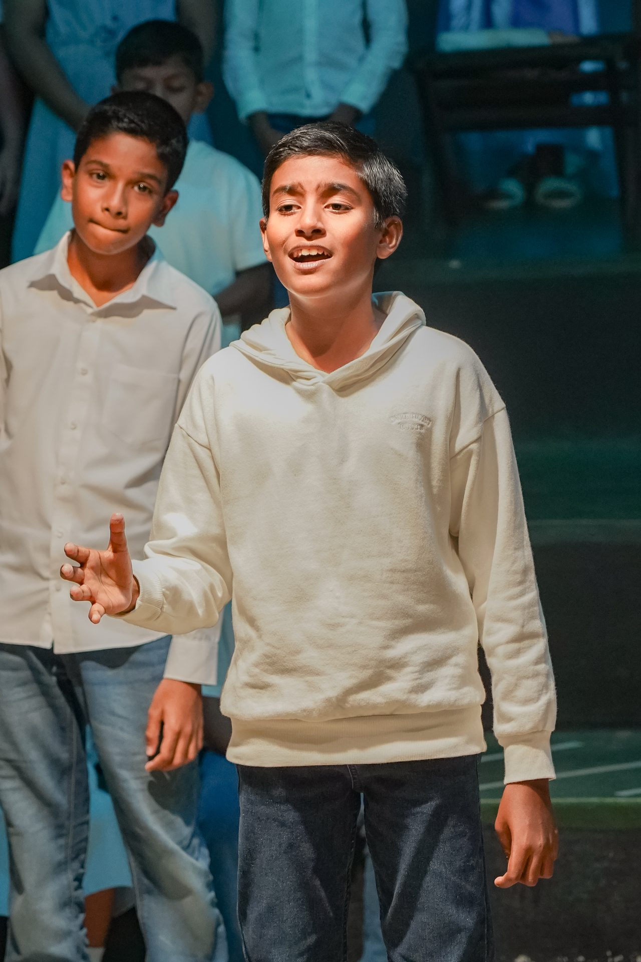 Two boys on stage: one singing passionately, the other listens attentively in a theater setting.