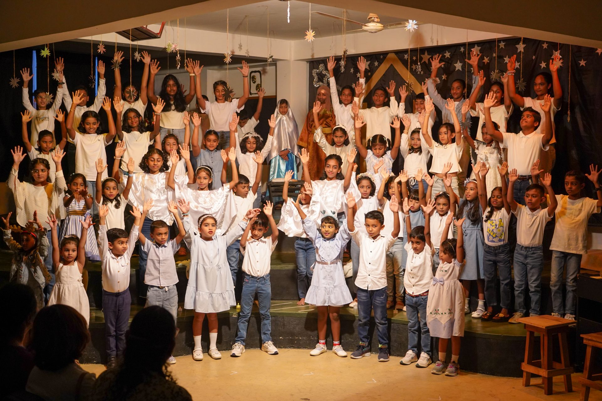 Children joyfully perform on stage, arranged in tiers, under warm lighting in pastel outfits.