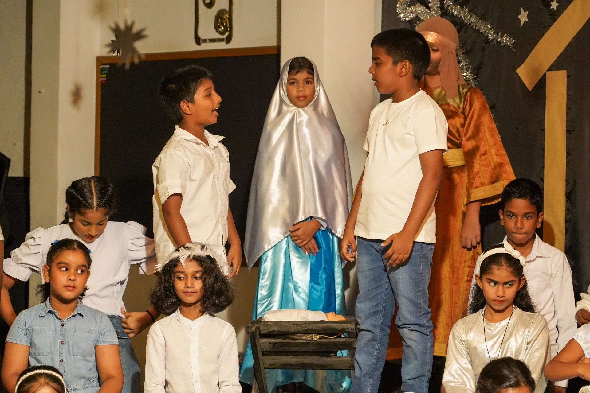 Children perform nativity play on stage with festive costumes and decorative backdrop.