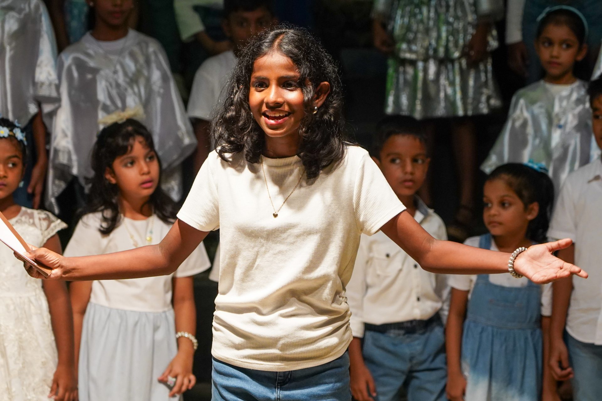 Curly-haired girl leads joyful childrens performance, arms open wide on stage.