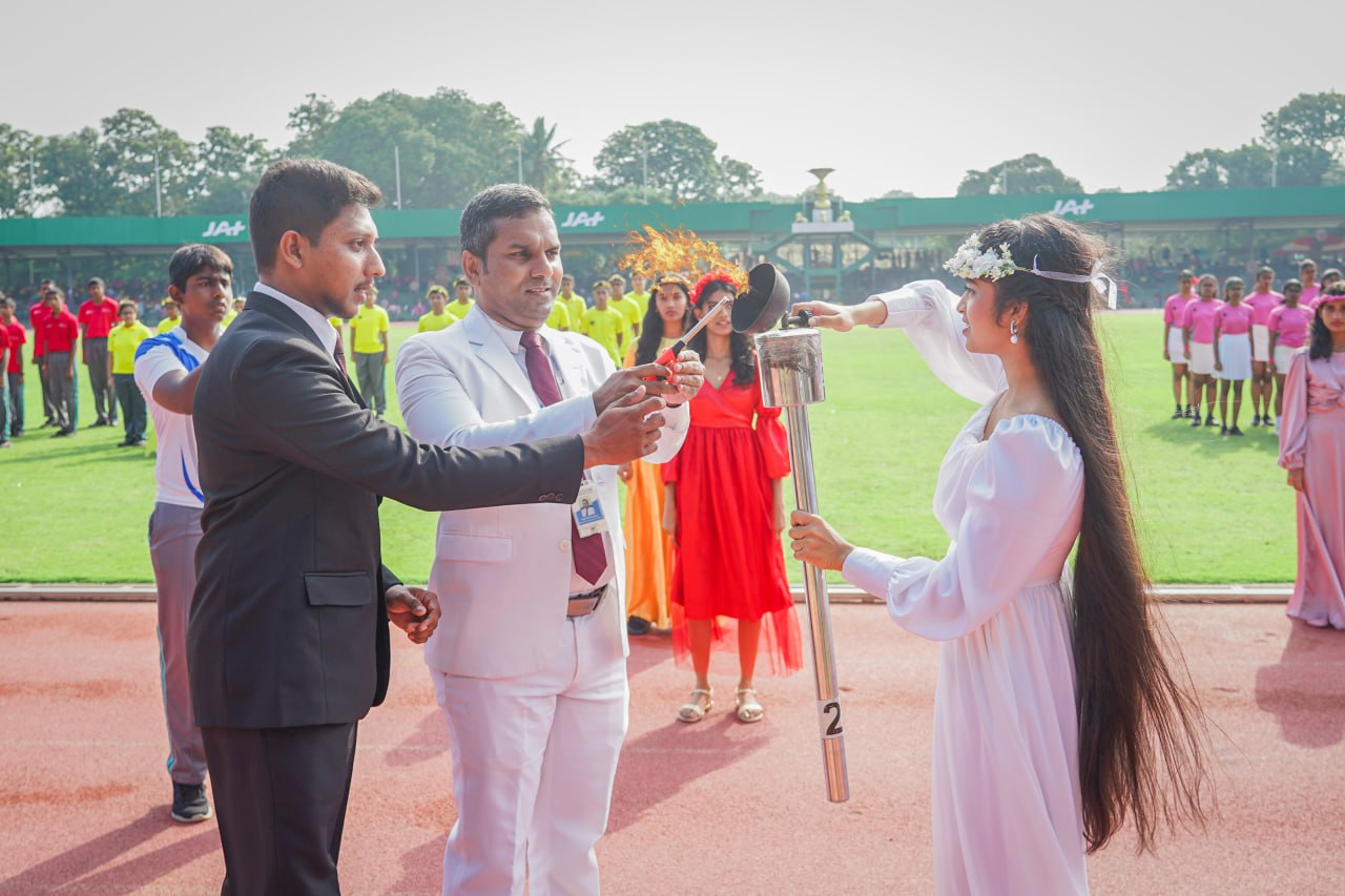 Woman torchbearer at outdoor ceremony, surrounded by colorful participants; grandstand labeled JAAF in background.