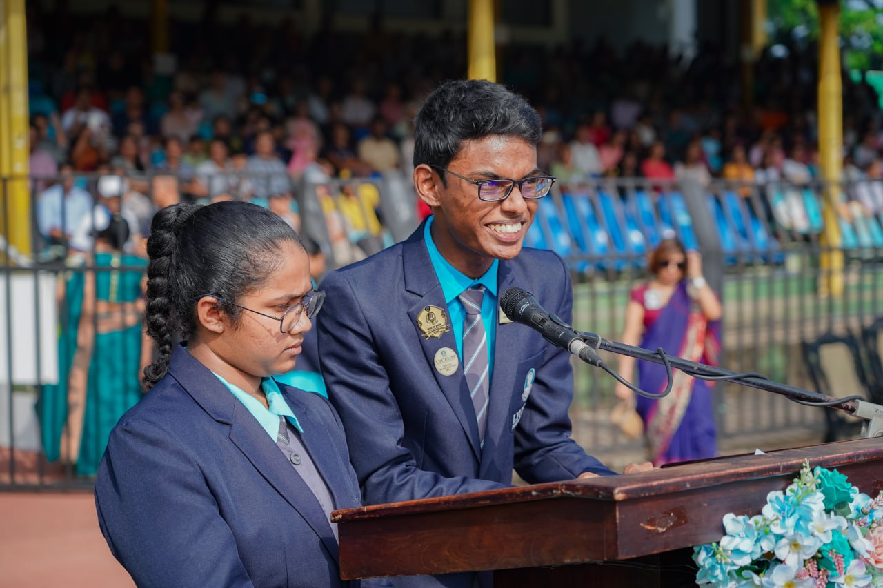Students speak confidently at school event podium, diverse audience in background under sunny skies.