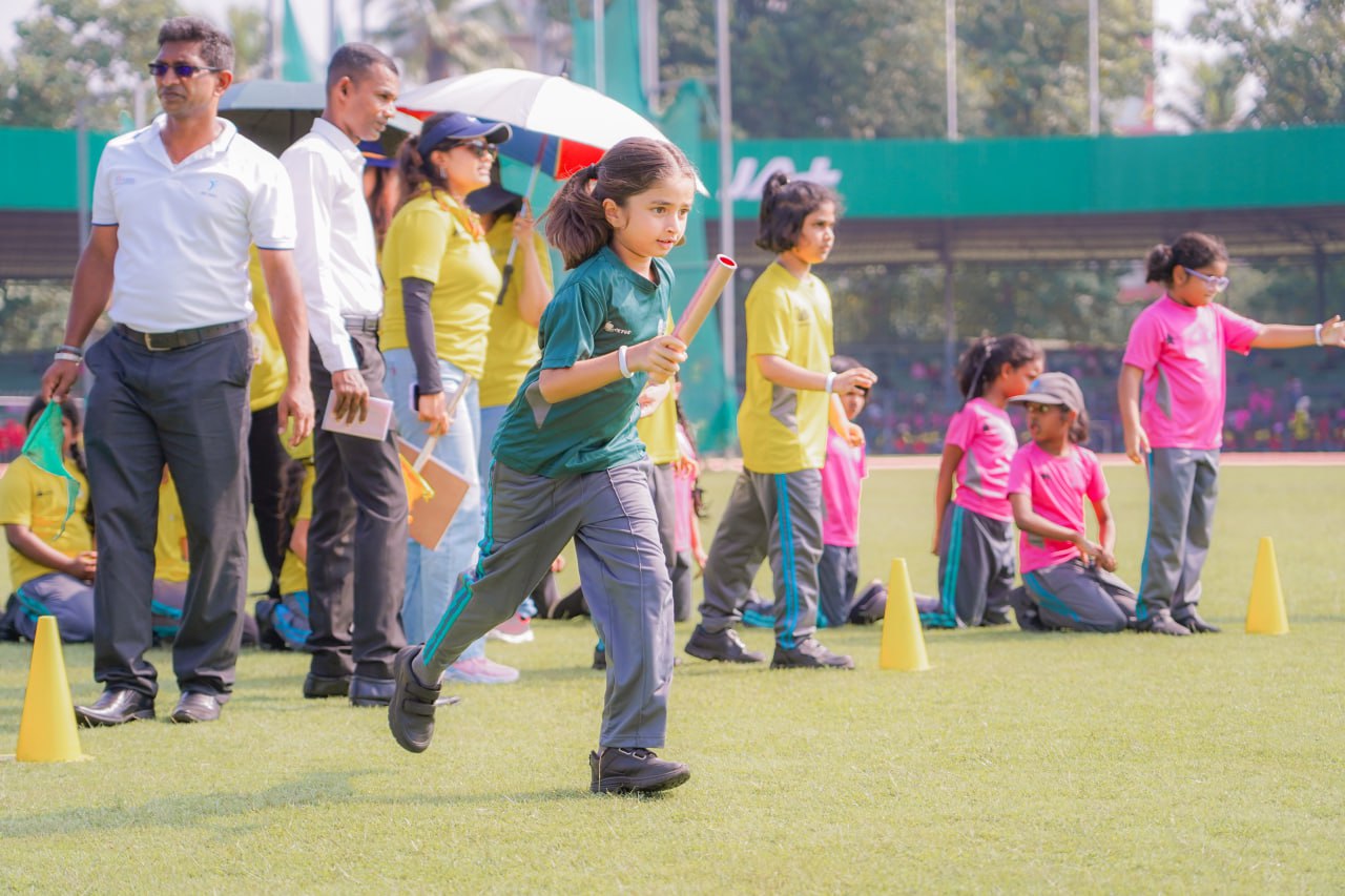 Girl runs in school relay race, carrying baton, with spectators and organizers on sunny sports day.