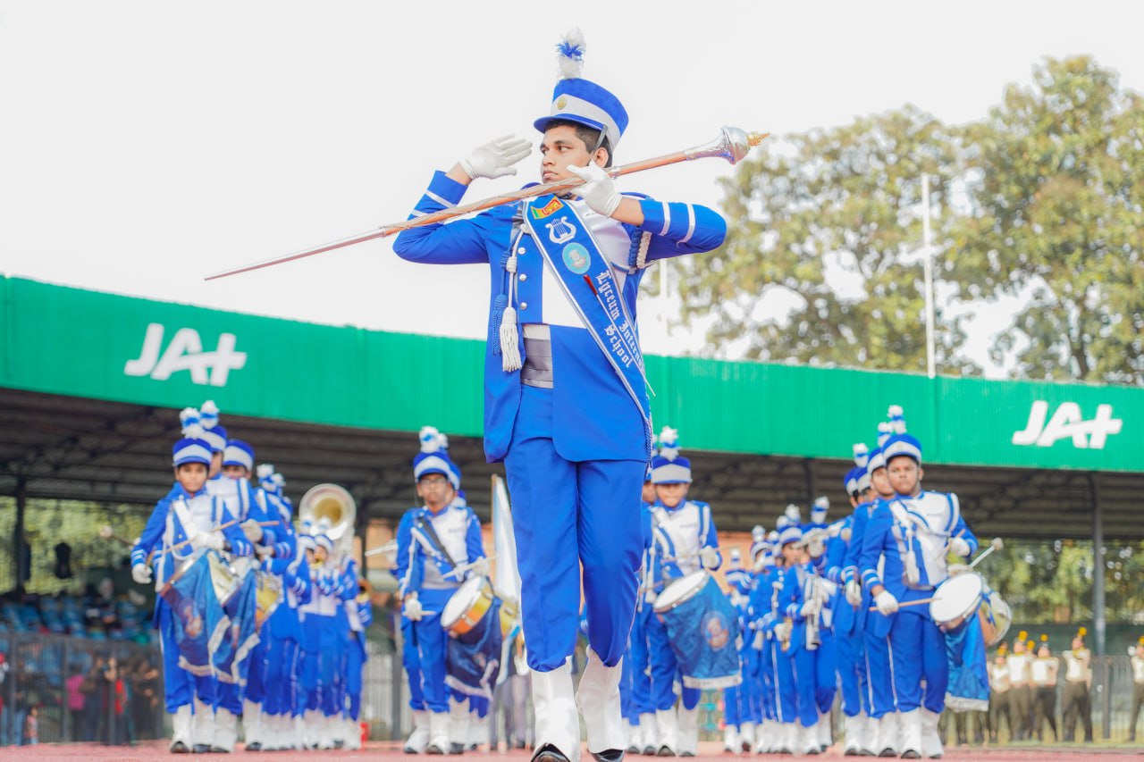Drum major leads synchronized blue marching band performance at lively outdoor JA+ event.
