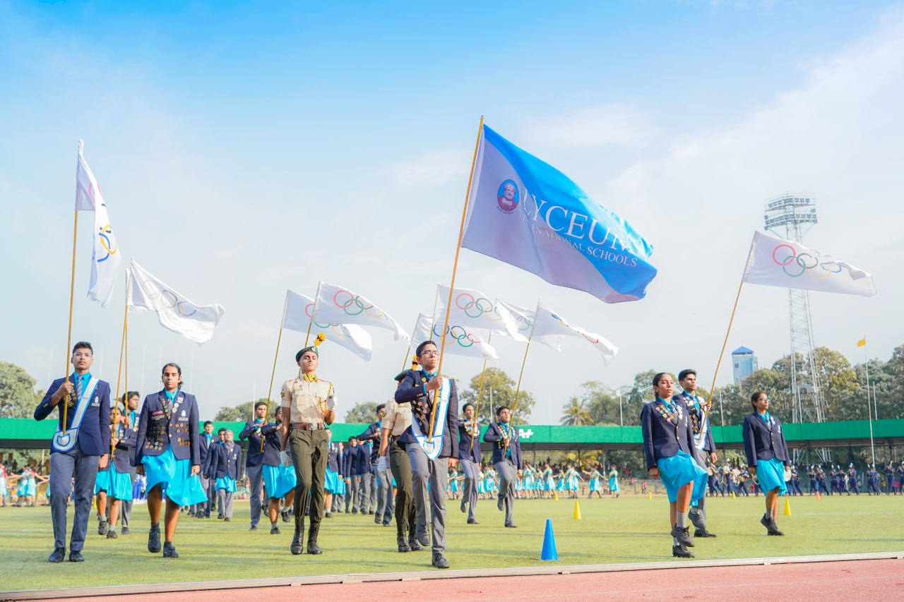 Students in a blue uniform parade holding ICEBO flags on a sunny school sports day.