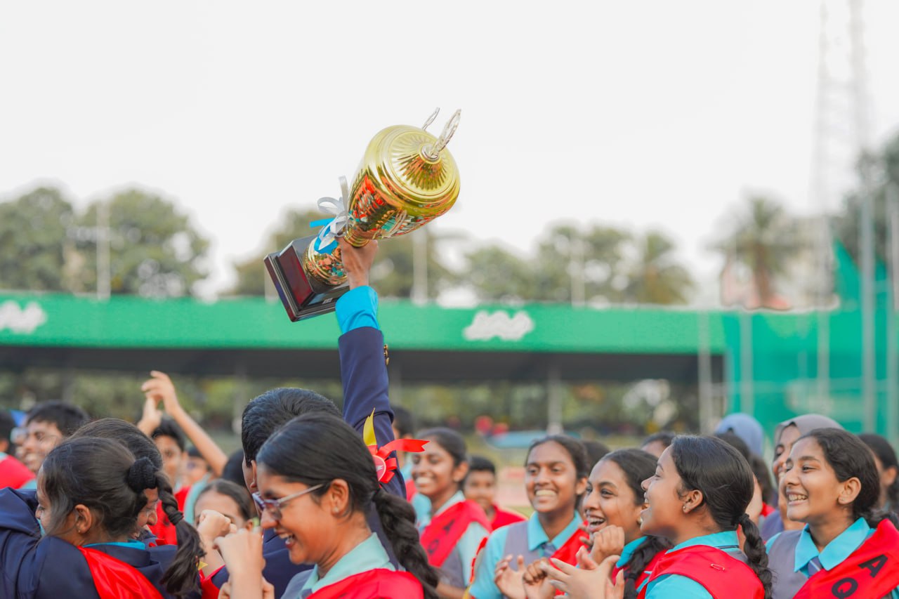 Students celebrate victory, holding golden trophy at school event, wearing blue and red uniforms.