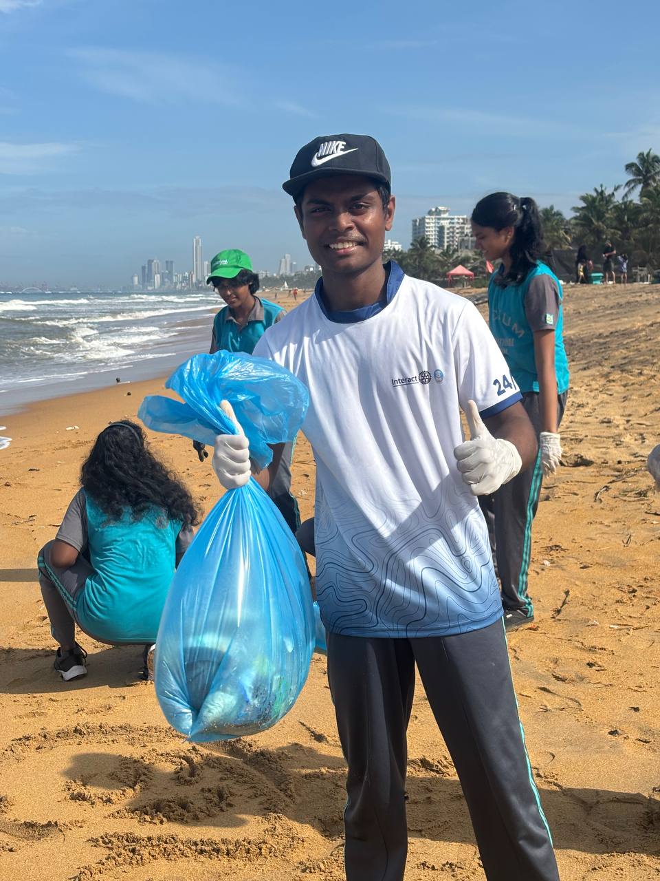 Smiling volunteer at beach cleanup with trash bag, city skyline, and palm trees in background.