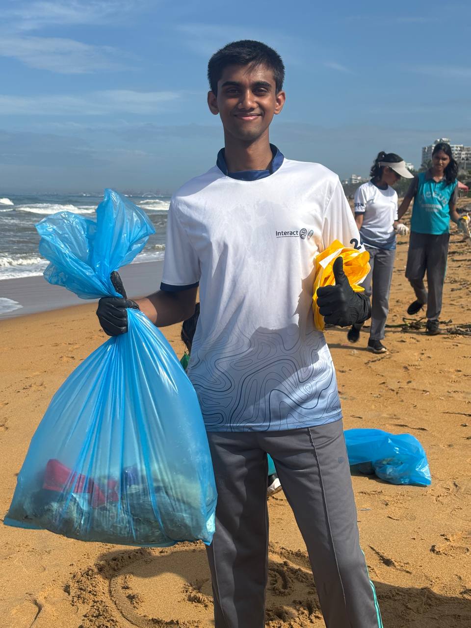 Young volunteer cleans beach, smiling with trash bag, amid sunny skies and community effort.