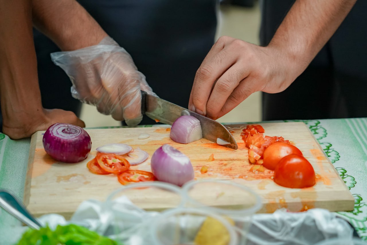 Gloved hand slicing fresh red onion amidst tomato slices on a wooden board.