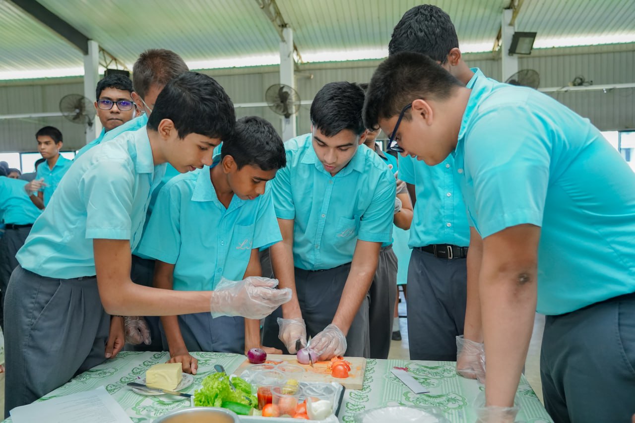 Schoolboys in teal uniforms collaboratively make sandwiches in a sunlit classroom.