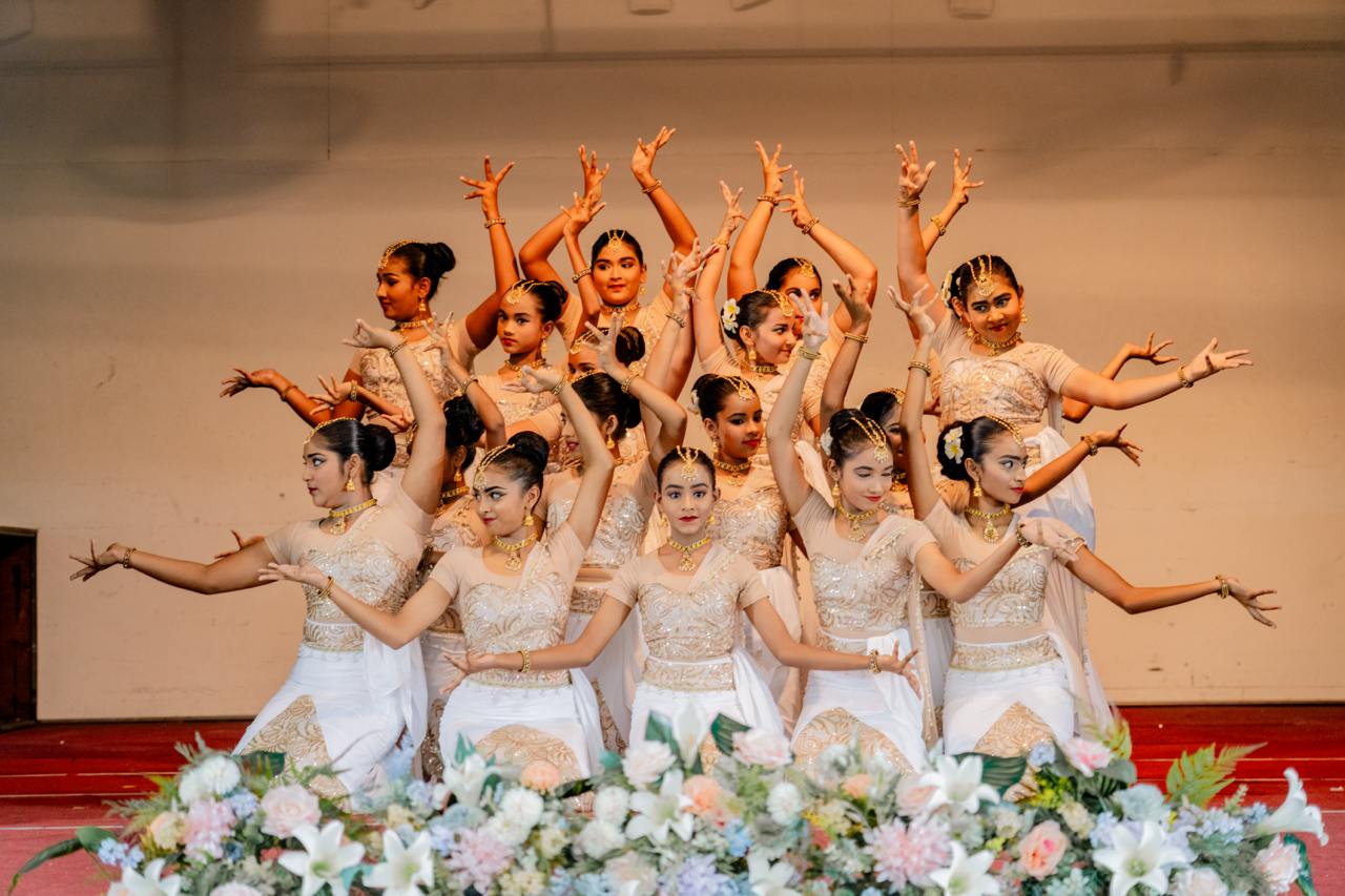 Traditional dancers in beige costumes perform synchronized choreography with expressive gestures on stage.