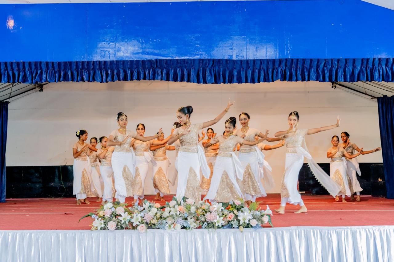 Elegant synchronized dancers in white and gold perform on a red-carpeted stage with floral decor.