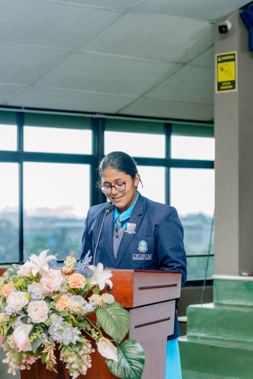 Student speaker at Lyceum School podium, floral decor, indoor natural light, COVID-19 precautions.