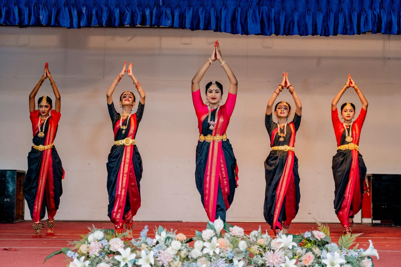 Five Indian dancers in traditional attire perform synchronized dance on a theatrical stage.