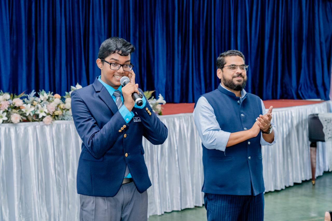 Formal event with speakers on stage, microphone, blue suits, and floral decorations.