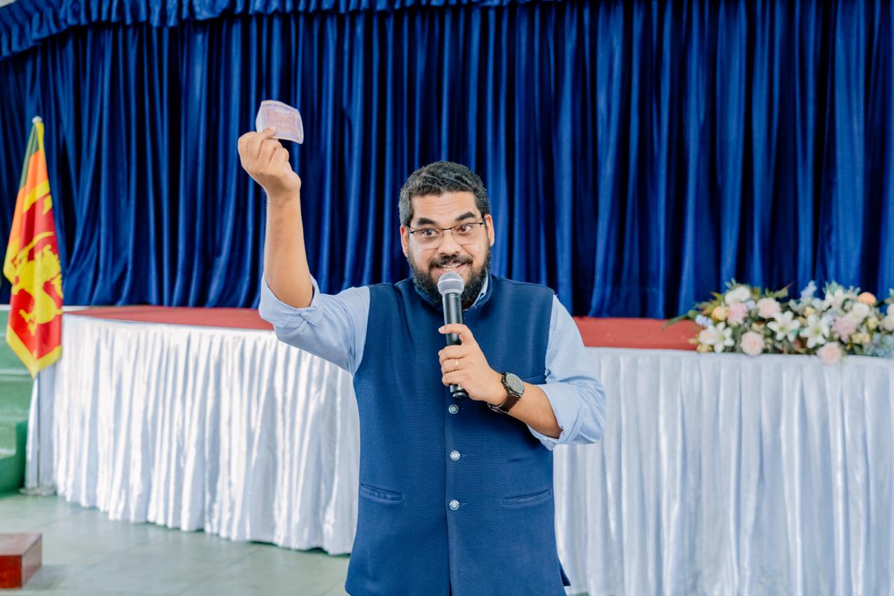 A man presents with card and microphone on stage, Sri Lankan flag in background.