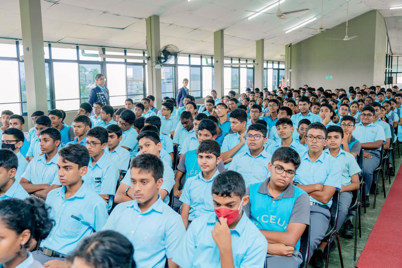Students in blue uniforms gather attentively in auditorium; one wears a red mask.