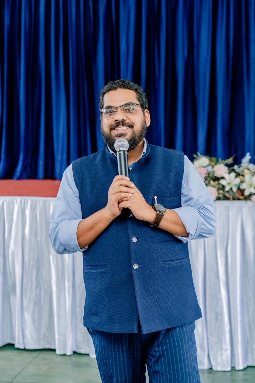 Speaker in Nehru jacket addresses audience at formal event with blue backdrop and flowers.