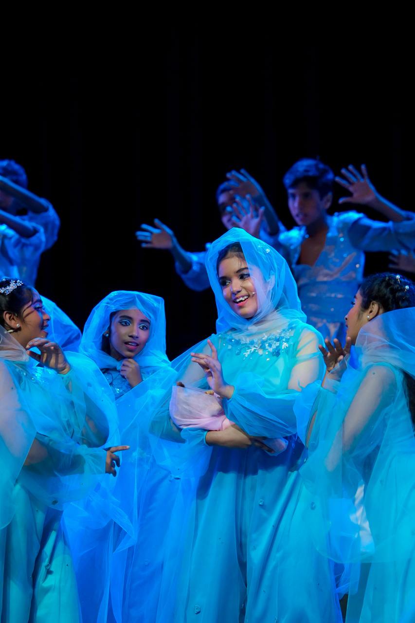 Performers in shimmering blue costumes captivate with dance and expressive gestures on a dark stage.
