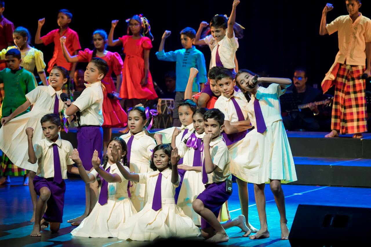 Children joyfully perform on stage in colorful outfits with live music accompaniment.