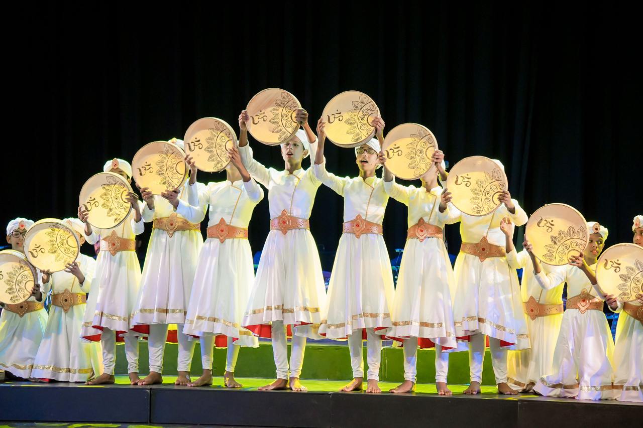 Traditional dancers in white with gold discs perform on a dimly lit stage.