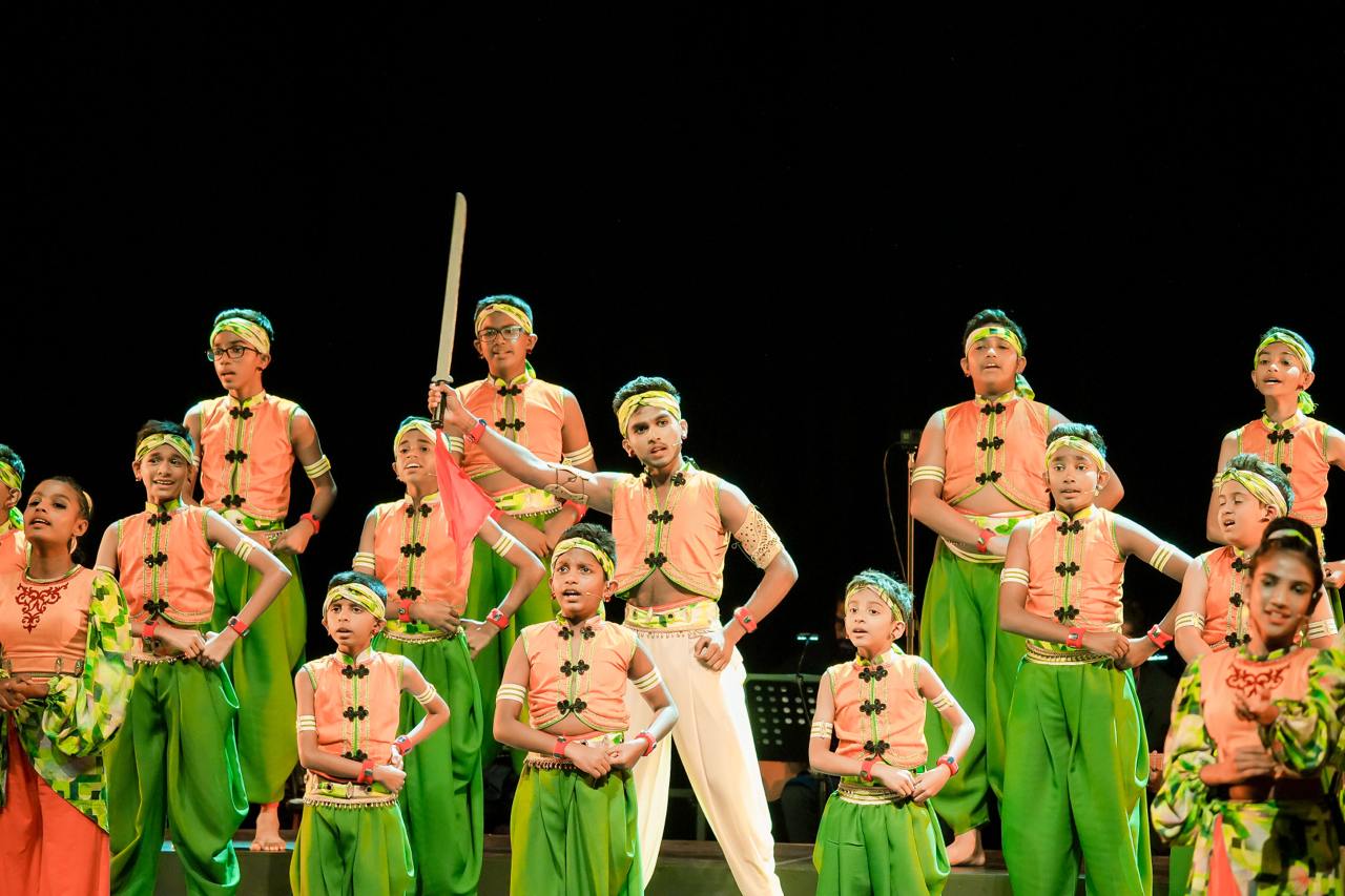 Young performers in vibrant green and peach costumes energetically dance on stage under bright lights.
