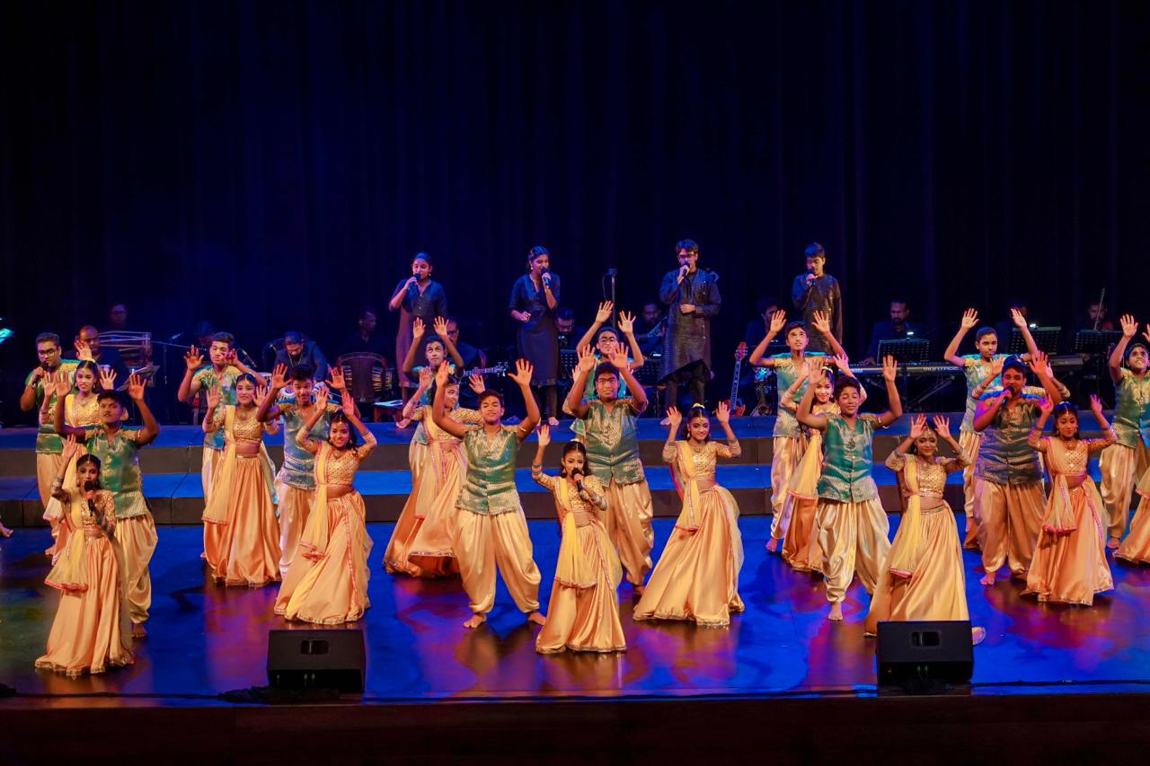 Energetic cultural dance with gold costumes, vibrant lighting, and live instrumental music on stage.