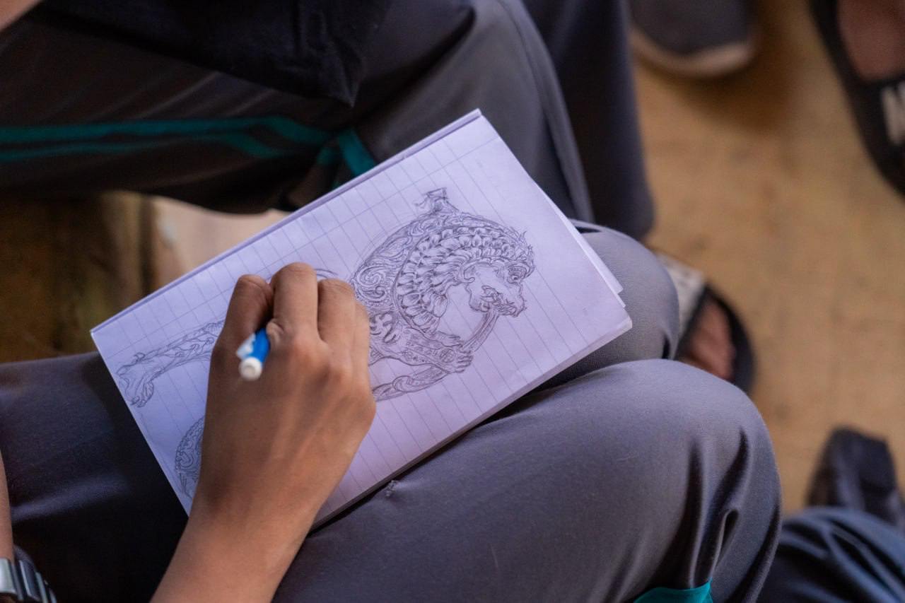 Person sketching intricate design on paper, seated casually outdoors, surrounded by natural light.