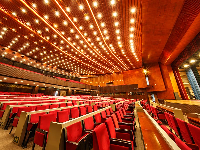 Elegant Conference Auditorium With Red Seats and Modern Lighting Grand auditorium with red seats, wooden ceiling, modern lighting, and stage for conferences or performances.