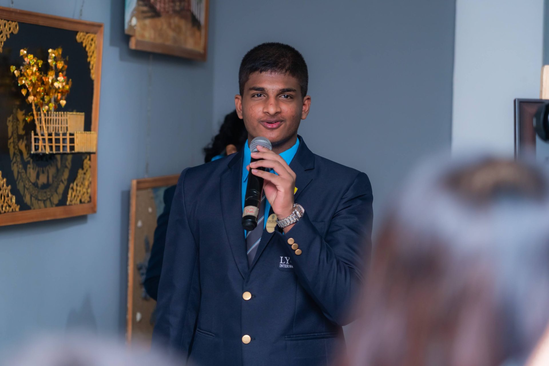 Confident student in navy blazer speaks with microphone at formal indoor school event.