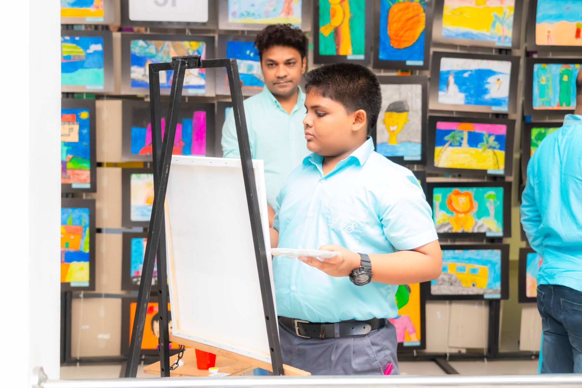 Young boy painting at kids art show, supported by mentor, with colorful children’s artwork displayed.