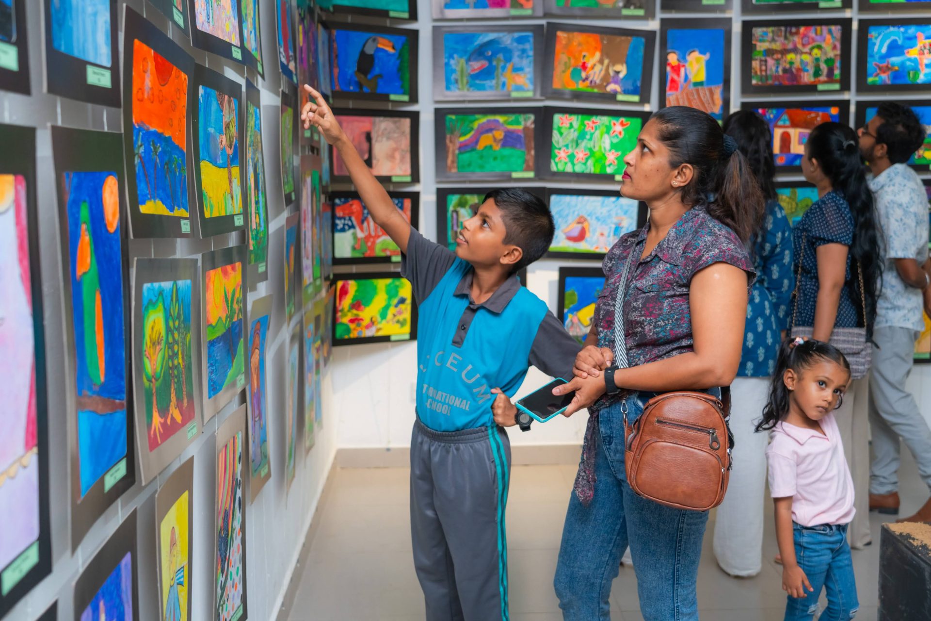 Family admires colorful childrens artwork at lively school art exhibition, young artist proudly sharing.