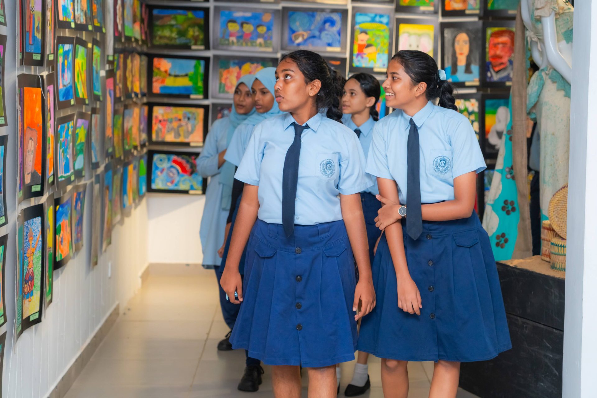 Schoolgirls in uniforms explore colorful student art and crafts at a bright school gallery exhibition.